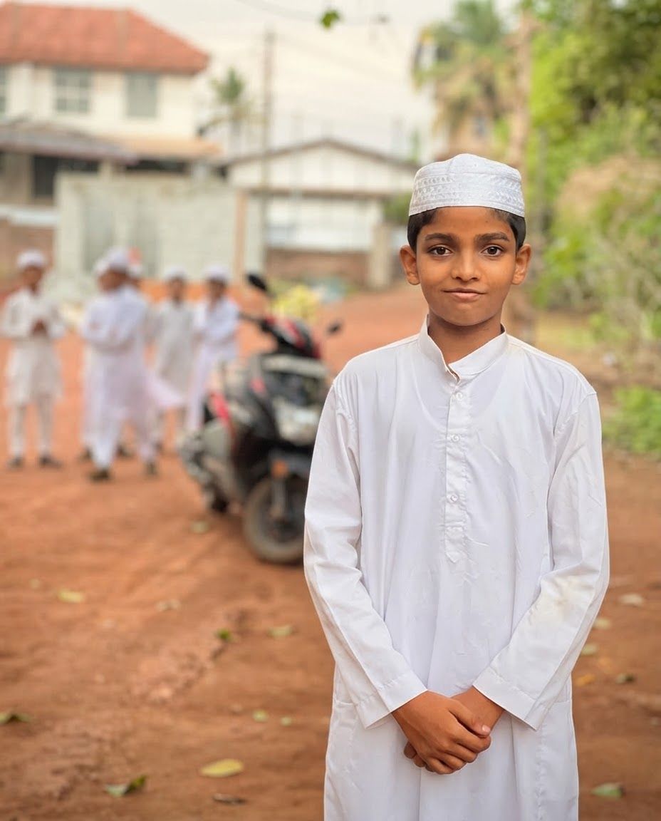 AI Design for Lifestyle product photograph of Traditional Youth Thobe being naturally used in a believable real-world setting. A young boy stands in the foreground wearing a white long-sleeved thobe and a matching white kufi cap. In the background, several other children in similar white attire are blurred on a dirt path with buildings and greenery visible. Human-scale context, warm natural light, editorial composition, shallow depth of field, product clearly visible and undistorted.
