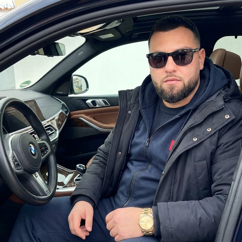 Szerkeszd meg ezt a képet úgy hogy van rajta 1 Hugoboss pulóver kabáttal. Benne ül a BMW-be. Kezén 1 arany rolex óra. És van rajta 1 napszemüveg. Az arca és a kép 100%-ban realisztikus legyen