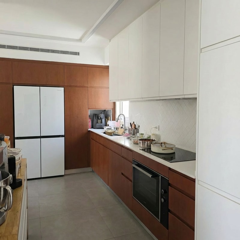 Change the refrigerator color to white.
• Reduce the refrigerator depth so it is fully flush with the cabinetry. Currently, the refrigerator protrudes 10 cm beyond the cabinet line; it should not protrude at all.
• On the opposite side, change the finish of the tall cabinets to a brown tone.
• Lower the height of the tall cabinets so they match the height of the tall cabinets in the refrigerator area, creating one consistent height for all tall cabinets in the kitchen.
• Continue the wall line above the cabinets so it wraps around them, creating a framed and cohesive look.