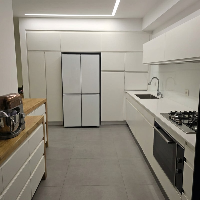 Edit the kitchen image so the refrigerator is flush and aligned in a straight line with the surrounding cabinets, not protruding. Change the refrigerator color to white. Remove all objects and clutter from the countertop, leaving it completely clean. Replace the electric cooktop with a gas cooktop featuring a stainless steel surface. Change the sink to a stainless steel sink. Keep the rest of the kitchen design, lighting, and proportions realistic and consistent with the original image