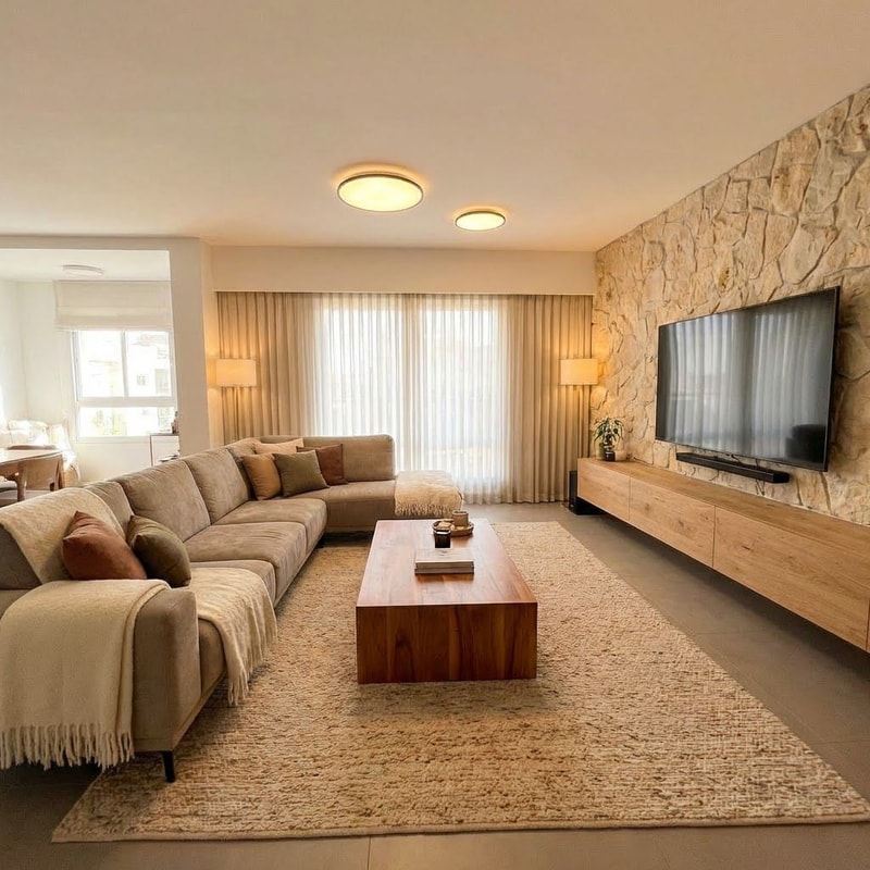 A cozy and elegant living room featuring a full-length sofa adorned with textured blankets and soft throw pillows, paired with a warm wood coffee table — Wide-angle interior shot with an expanded field of view to show the entire sofa and the full expanse of the large window area, A harmonious palette of warm earth tones, rich wood grains, and creamy neutrals, Soft ambient lighting creating a warm, inviting, and comfortable atmosphere, Photorealistic interior design visualization with a focus on natural textures and elegant materials, Ensure the entire sofa and coffee table are fully visible within the frame without cropping; avoid cold or harsh lighting, A bright residential interior featuring a large window that provides a soft backdrop, grounded by a textured, warm-toned area rug.