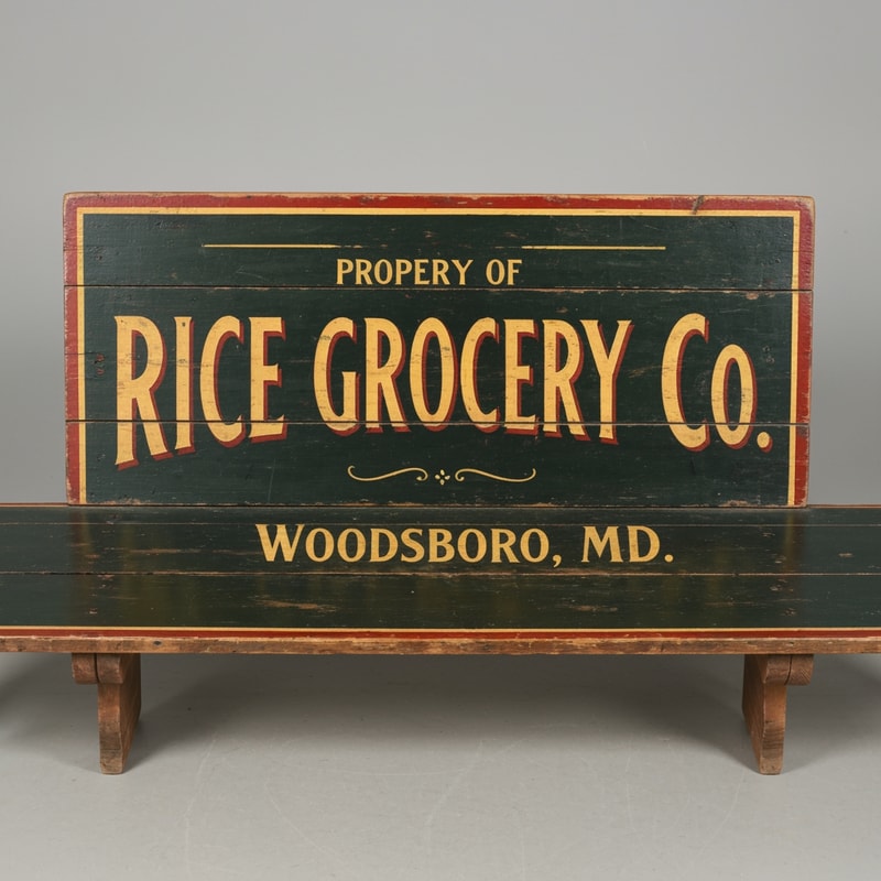 A vintage wooden bench designed like an old general store advertising sign, viewed straight from the front (perfect frontal view, no perspective distortion). The top surface is flat and clearly visible like a signboard. It has hand-painted, slightly worn lettering in a classic 19th-century style. The main text reads “Rice Grocery Co.” in large, bold, warm yellow and red lettering. Above it in smaller text: “Property of”. Below it: “Woodsboro, MD.” The wood is aged, slightly weathered with visible grain and subtle paint wear. Colors are dark green, black, and rustic tones. The bench has simple wooden legs visible underneath. Lighting is soft and natural, background neutral and unobtrusive.
